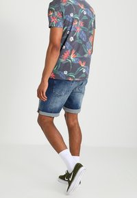 Hombre que lleva una camisa de manga corta con estampado floral, pantalones cortos de denim enrollados, calcetines blancos y zapatillas deportivas verdes, de pie contra un fondo sencillo.