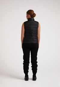 Gilet nero imbottito con colletto alto e cuciture verticali, indossato sopra dei joggers neri. Il gilet ha una texture liscia e lucida ed è senza maniche.