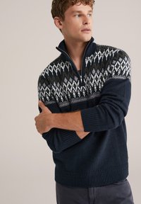 Pulover tricotat cu fermoar, de culoare bleumarin, cu un model Fair Isle în verde și alb. Dispune de manșete și tiv ribbati. Materialul pare moale și texturat.