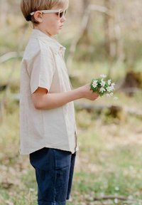 Kortärmad beige skjorta, marinblå shorts, håller vita blommor, bär vita solglasögon, utomhus med suddig grönska i bakgrunden.