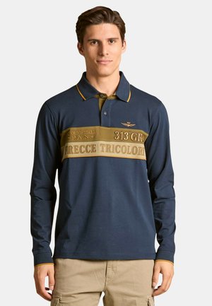 Uomo che indossa un polo a maniche lunghe blu navy con una striscia senape e il testo "Frecce Tricolori", abbinato a pantaloni cargo beige, in piedi contro uno sfondo neutro.
