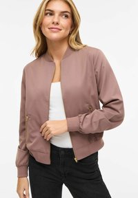 Tânără cu părul blond, purtând o geacă bomber mov cu fermoar peste un top alb și pantaloni negri, pe un fundal alb simplu.