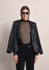 Schwarze Satin-Blazer mit breiten Revers, getragen über einem durchsichtigen, strukturierten braunen Oberteil. Der Look umfasst hochtaillierte schwarze Satin-Hosen und Sonnenbrillen.