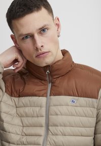 Polstret zip-jakke med en brun overdel og en lys beige underdel, med en quiltet struktur og et lille logo på brystet.