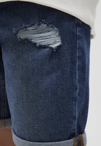 Short en jean bleu foncé avec une pièce déchirée laissant apparaître un tissu plus clair. Les revers sont roulés et les coutures sont marron contrastées.