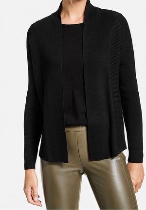 Kobieta ubraną w czarny prążkowany kardigan z otwartym frontem nałożony na czarną bluzkę, w połączeniu z oliwkowozielonymi skórzanymi spodniami.