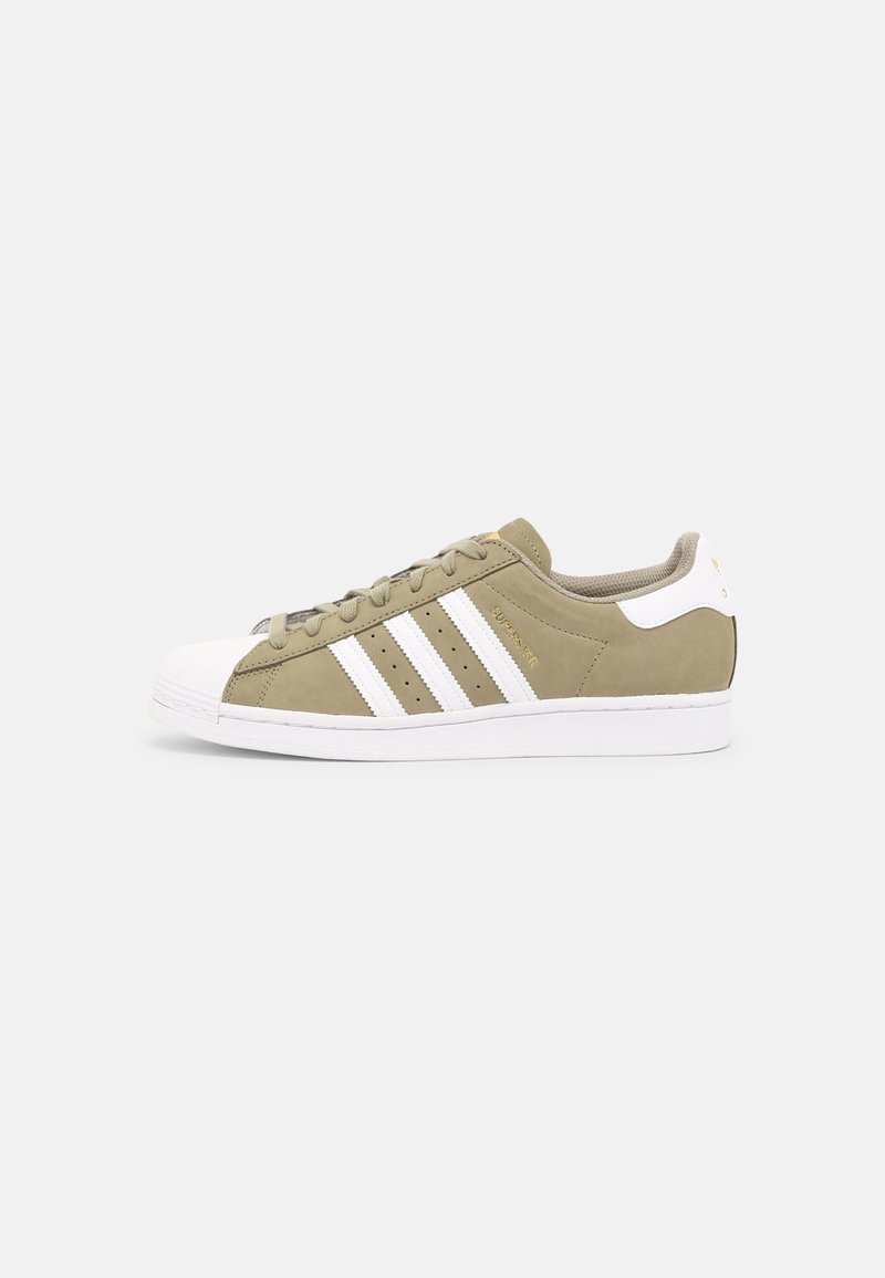 Beige Adidas Superstar-sneaker med vit snabelfot, vita ränder och vit sula visad i sidoprofil på en neutral bakgrund.