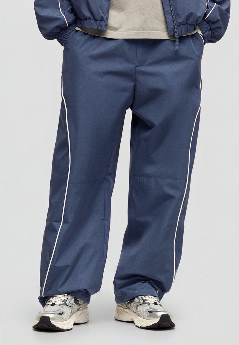 Personne portant un pantalon de survêtement bleu ample avec passepoil blanc, un t-shirt gris, une veste bleue assortie et des baskets blanches, debout sur un fond clair.