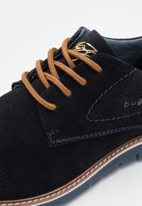 Chaussure en daim bleu marine avec lacets marron, coutures bleues, panneau latéral perforé et semelle marron crantée.