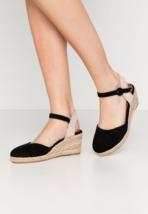 Ben iført sorte wedges med espadrille-sandaler i ruskind med beige flettede hælremme og ankelspænder på en lys baggrund.
