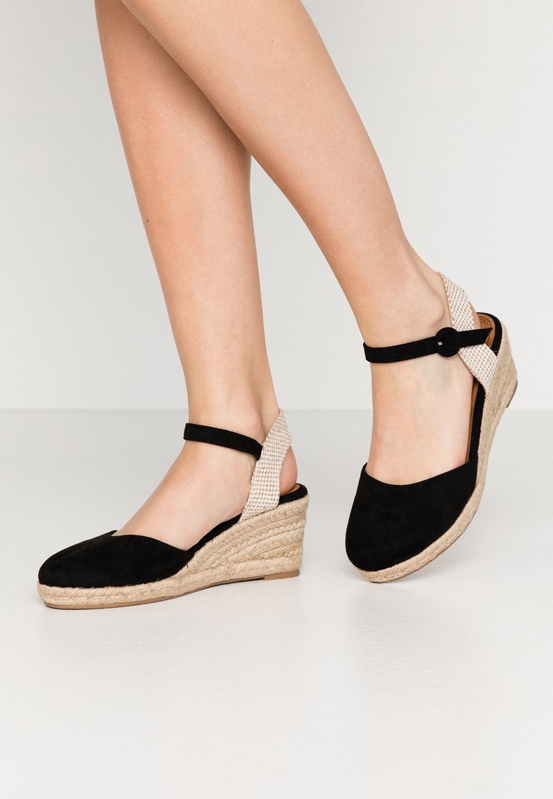 Jambes portant des sandales compensées espadrilles en suède noir avec des brides tissées beige autour du talon et des boucles à la cheville sur un fond clair.
