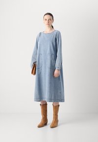 Jeanskleid mit langen Ärmeln, rundem Ausschnitt, zwei großen Taschen und gerafften Bündchen. Hellblaue Farbe mit sichtbaren Nahtdetails. Braune Lederstiefel.