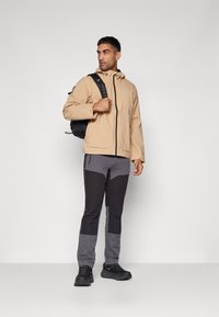 Hellbeige wasserdichte Jacke mit Kapuze, schwarzen Reißverschlüssen und zwei Fronttaschen. Kombiniert mit grauen und schwarzen engen Hosen und schwarzen Schuhen.