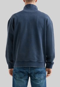 Marineblauer Pullover mit hohem Kragen, lockerer Passform, geripptem Saum und Bündchen sowie einer dezenten strukturierten Oberfläche. Getragen mit blauen Jeans.