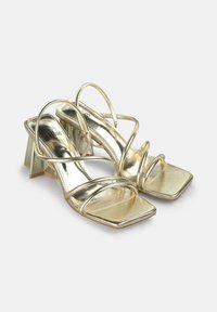 Goldene metallische Absatzsandalen mit mehreren schlanken Riemen, einer quadratischen Zehenpartie und einem geometrischen Blockabsatz mit transparentem Akzent.