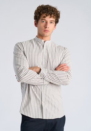 Ung mand med krøllet hår iført en grå og hvid vertikalt striped skjorte, stående med armene krydset mod en ensartet lys baggrund.