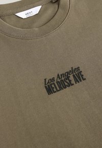 Olivengrøn bomuldscrewneck t-shirt med ribbet krave. Har sort broderet tekst: "Los Angeles Melrose Ave." Glat tekstur.