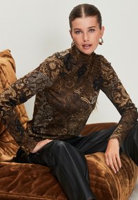 Brown turtleneck top featuring intricate lace and floral patterns, with long sleeves. Paired with black, shiny, pleated trousers.