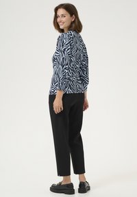 Sieviete, kas smaida pāri plecam, nēsājot zilā un melnā zebras raksta blūzi, melnas īsas bikses un melnas loafer kurpes uz baltā fona.