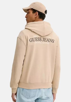Jonge man met een beige hoodie en bijpassende pet, zijwaarts kijkend met de tekst "Guess Jeans" zichtbaar op de achterkant van de hoodie.
