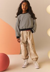 Wzorzysty sweter w czarno-białe paski z elastycznym ściągaczem na dole, beżowe spodnie cargo z elastycznymi mankietami, białe sneakersy typu high-top, czerwona spinka do włosów.