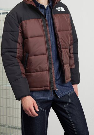 Man som bär en brun och svart North Face-puffjacka över en blå denimskjorta och mörka jeans med vit söm.