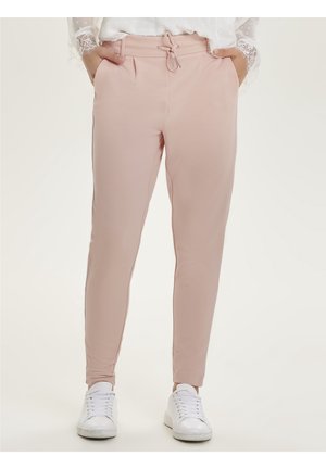 Femme portant un pantalon tapered rose clair avec taille à cordon, poches latérales et baskets blanches, se tenant avec les mains dans les poches.
