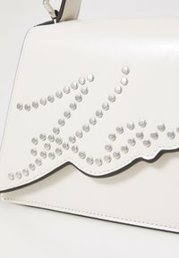 White leather handbag with silver studded pattern, wavy edge design, and black stitching. Features a shoulder strap for carrying.