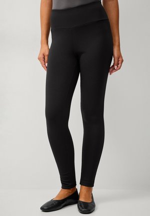 Kvinde iført sorte tætsiddende leggings og sorte balletsko, der står mod en ensfarvet baggrund.