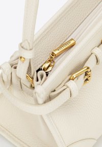 Crème leren handtas met een gek textured afwerking, ritsluiting en gouden hardware accenten, waaronder een gelabelde rits trekker.