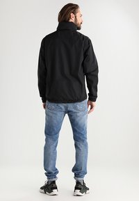 Veste noire en nylon avec un col montant, des poignets élastiques et un ourlet, portée sur un jean bleu clair et des chaussures de sport noires, vue de dos.