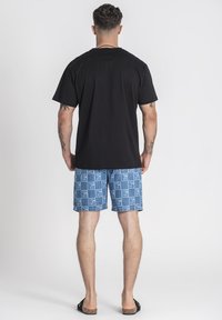 Černé tričko, ležérní střih; světle modré šortky s opakovaným vzorem loga; ploché sandály; pohled zezadu na jednotném pozadí.