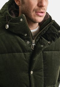 Dunkelgrüne Cordjacke mit gestepptem Design, ausgestattet mit einem Reißverschluss, Druckknöpfen, einem hohen Kragen und einer verstellbaren Kapuze.