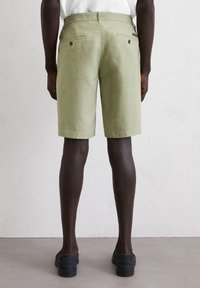 Light green linen shorts with a straight cut, featuring two back pockets with brown buttons, paired with dark shoes on a neutral background.