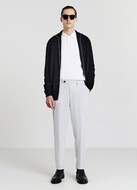 Schwarze Reißverschluss-Strickjacke, weißes Polo-Shirt, hellgraue maßgeschneiderte Hose, schwarze Stiefeletten und schwarze Sonnenbrille. Glatter Stoff und strukturierte Passform.