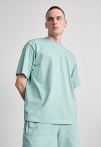 Tricou scurt cu mânecă în verde mentă, confecționat dintr-un material bumbac moale. Croială lejeră cu decolteu rotund și detaliu subtil al logo-ului pe piept. Piesă inferioară asortată.