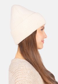 Bonnet en tricot blanc à côtes avec un sommet pointu, motif texturé et bord retroussé, porté par une personne ayant de longs cheveux bruns.