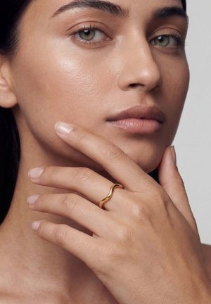 Femeie atingându-și bărbia cu degetele, purtând un inel auriu neted și unduitor pe degetul mijlociu, pe un fundal neutru.