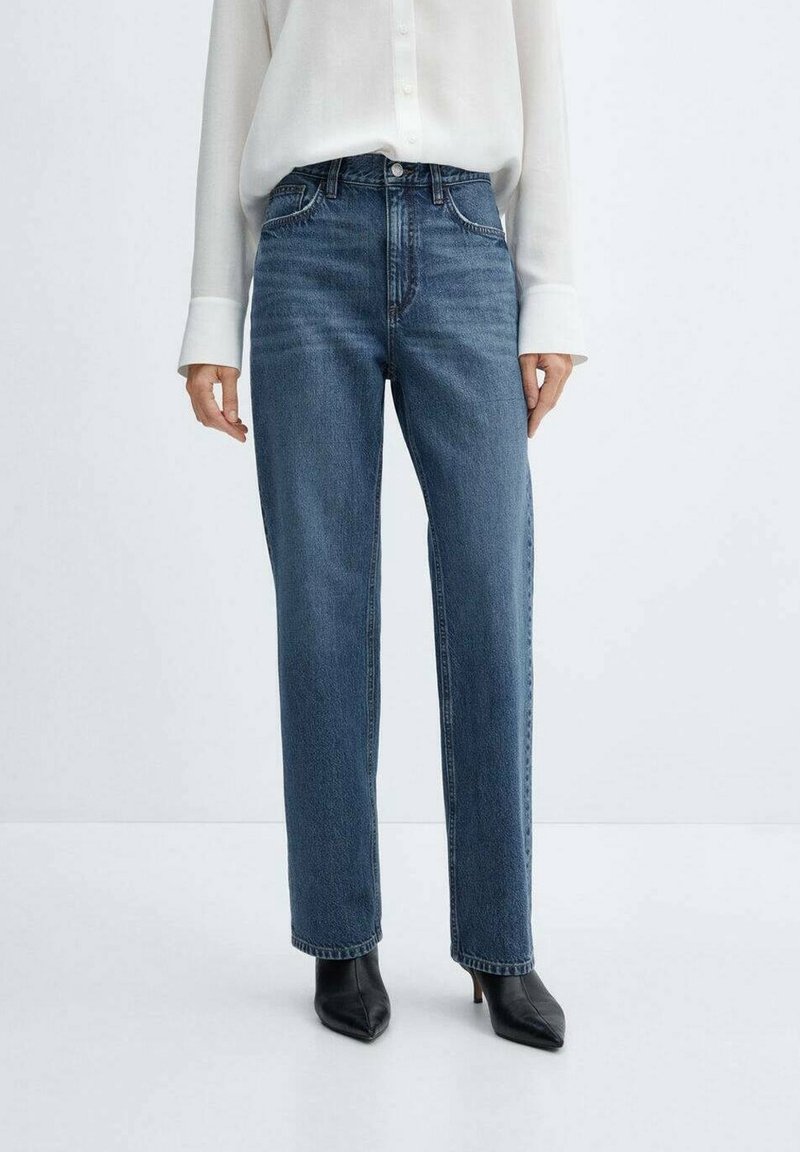 Jeans in denim blu a gamba dritta con vita alta, cuciture accentuate e tasche frontali, abbinati a una camicia bianca con bottoni e tacchi neri.