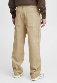 Beige cargo bukser lavet af en bomuldsblanding med en løs pasform, der har flere lommer og syede detaljer. Båret med hvide sneakers.