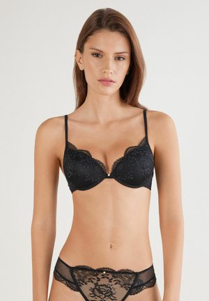 Reggiseno in pizzo nero con coppe imbottite, bordi ondulati e spalline regolabili. Abbinato a un perizoma in pizzo nero con motivi floreali.