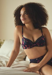 Sieviete ar lokainiem matiem un pūkainēm sejas ādām sēž uz gultas, ģērbta tumši zila ziedu krāsā krūšturī un saskanīgās augstās biksītēs, skatoties uz sāniem.