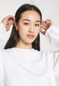 Sweatshirt branco com mangas franzidas, vestido por uma pessoa. Textura de tecido suave, design simples e estilo minimalista, com maquilhagem natural e joias.