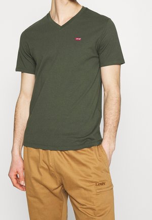 Mężczyzna w oliwkowozielonej koszulce V-neck marki Levi’s oraz beżowych spodniach typu cargo z logo Levi’s, stojący z jedną ręką w kieszeni na jednolitym tle.