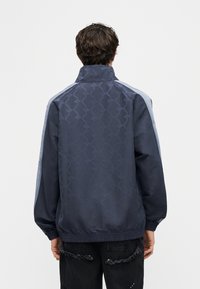 Νεαρός με την πλάτη στην κάμερα, φοράει σκούρο μπλε μπουφάν με ανοιχτό γκρι ρίγες στα μανίκια και μαύρα σκισμένα τζιν.