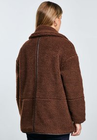 Brauner, flauschiger Fleece-Jacke mit entspannter Passform, hohem Kragen und kontrastierender brauner Borte entlang der Rückennaht.