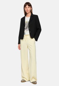 Schwarzer Blazer mit tailliertem Schnitt, Knopfverschluss und zwei vorderen Taschen, getragen über einem weißen T-Shirt mit Grafikdruck und hellgelben Hosen.