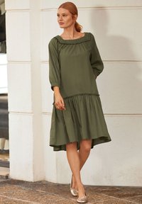 Olivengrünes Kleid mit gerafftem Ausschnitt, Dreiviertelärmeln und gestuftem Saum. Glattes Material mit subtiler glänzender Oberfläche und seitlichen Taschen.