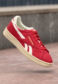 Sneaker in tela rossa con accenti bianchi, punta rotonda e suola in gomma beige. Presenta un logo sul lato e lacci per la chiusura.