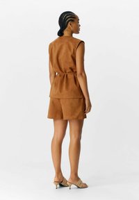 Haut sans manches en suede brun assorti à un short, avec une taille ceinturée et une texture lisse. Porté avec des sandales à talons beiges clairs.